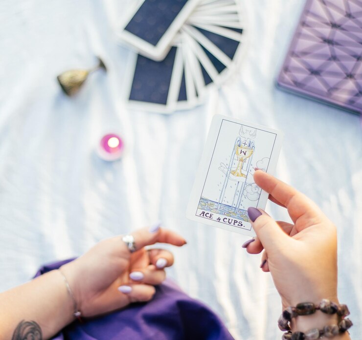 Une main féminine avec des ongles violets tient une carte de tarot nommée Ace of Cups sur une surface blanche avec un cahier et une bougie. Vue de dessus. Minsk, Biélorussie - 28/07/2021
