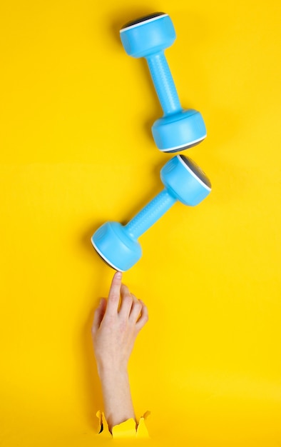 Une main féminine prend un haltère en plastique bleu à travers du papier jaune déchiré. Concept de sport minimaliste