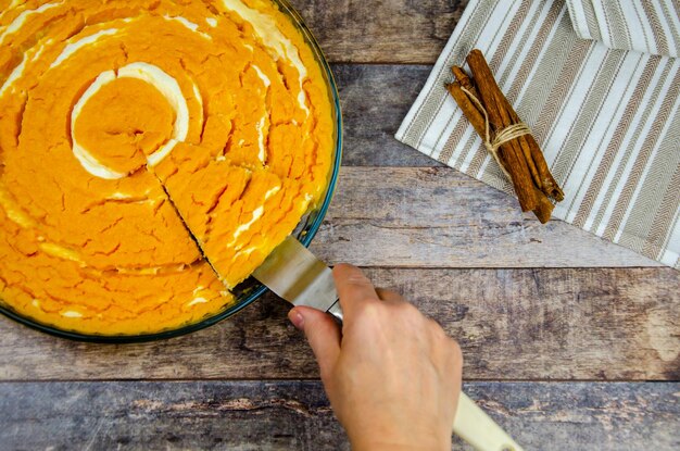 La main féminine prend un morceau coupé de casserole de caillé de citrouille avec une spatule. Cheesecake à la citrouille, table en bois