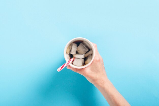 Main féminine prend ou tient un verre avec un café glacé rafraîchissant sur une surface bleue. Concept de chaleur, soif, cola avec de la glace. Mise à plat, vue de dessus