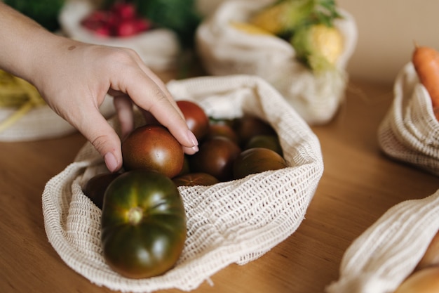 Une main féminine prend une tomate dans un sac d'épicerie en toile. Légumes dans des sacs en coton écologique réutilisables sur table en bois. Concept d'achat zéro déchet. Articles sans plastique