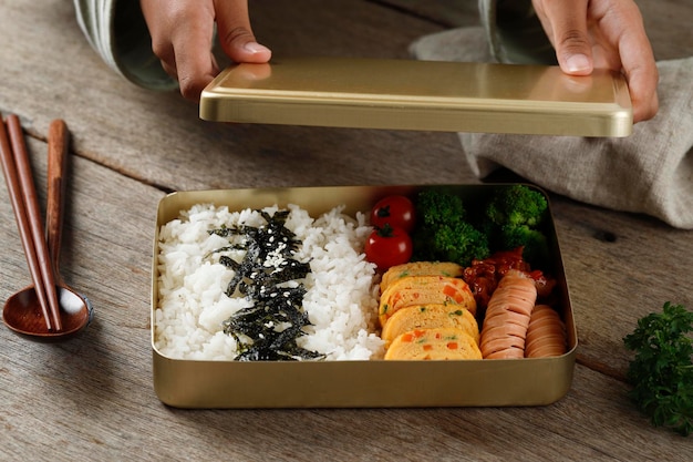 Main féminine préparant Dosirak ou boîte à bento coréen servie sur une boîte à lunch en métal doré pour garder les aliments au chaud