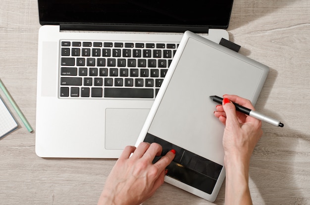 Main féminine avec un stylet sur une tablette graphique, ordinateur portable ouvert sur une table lumineuse, vue de dessus