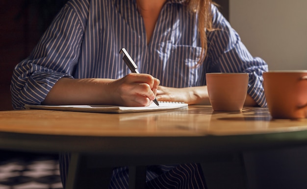 Main féminine avec un stylo prenant des notes par écrit des plans dans un journal sur une table en bois avec des tasses à café