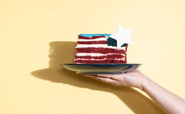 Main féminine tenant une assiette avec un morceau de gâteau sous la forme du drapeau des États-Unis sur fond jaune, célébrant le jour de l'indépendance, en gros plan.