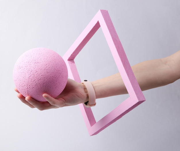 Main féminine tenant le ballon à travers un cadre rose sur fond gris Idée fraîche de minimalisme