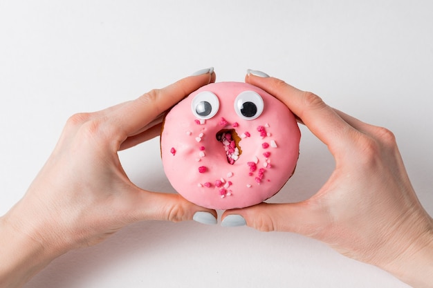 Main féminine tenant un beignet rose mignon avec une drôle de tête et des yeux. Donut sur blanc.