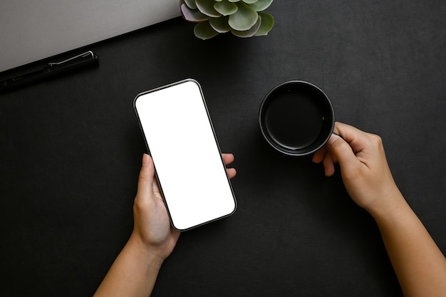 Une main féminine tenant une maquette de smartphone et une tasse de café noire sur une table noire moderne