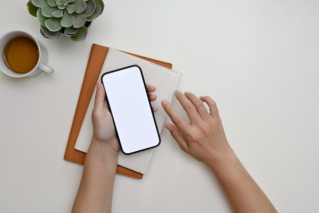 Photo une main féminine tenant une maquette de smartphone sur une vue de dessus de bureau blanche minimale