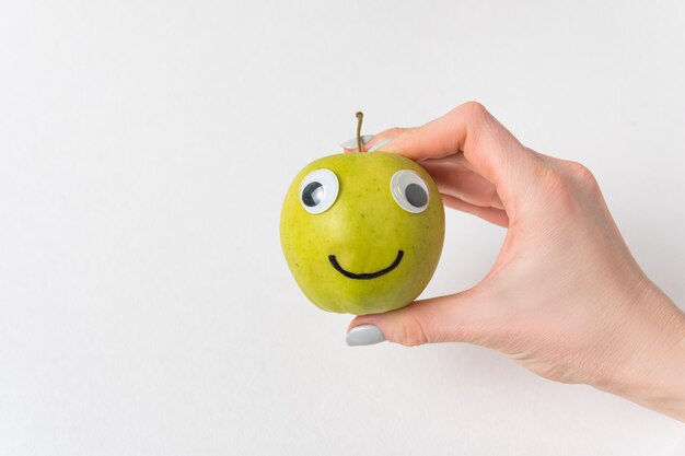 Main féminine tenant le petit sourire d'Apple avec les yeux écarquillés. Fond blanc. Concept de régime et de perte de poids.