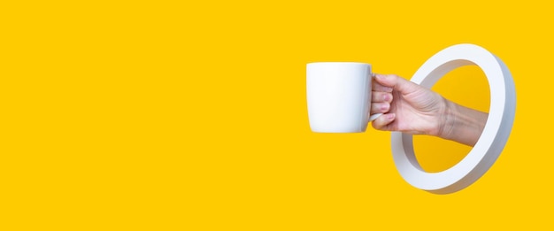 Main féminine tenant une tasse blanche dans un trou rond sur fond jaune. Bannière.