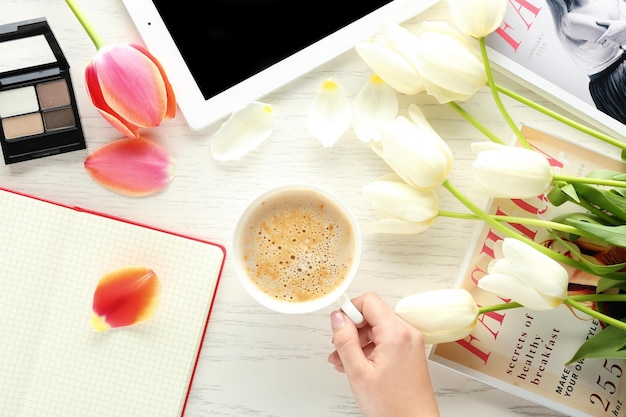 Main féminine tenant une tasse de café sur la table avec des magazines de cahier de fleurs et des cosmétiques