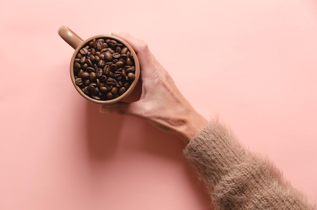 Main féminine tenant une tasse de grains de café sur fond rose