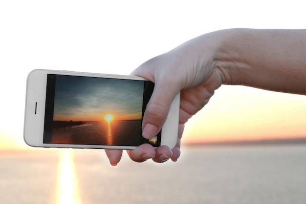 Main féminine tenant un téléphone intelligent et prenant une photo du coucher du soleil