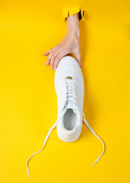 Une main féminine tient des baskets blanches à travers du papier jaune déchiré. Concept de mode créative minimaliste