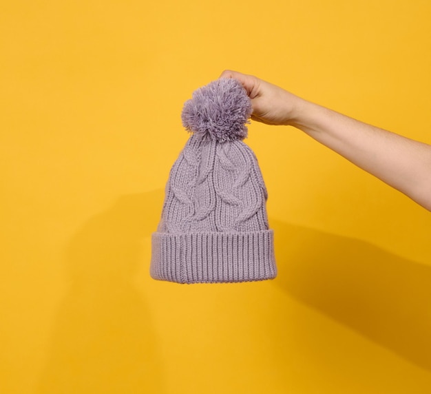 La main féminine tient un bonnet tricoté avec un pompon sur fond jaune, des vêtements chauds et confortables