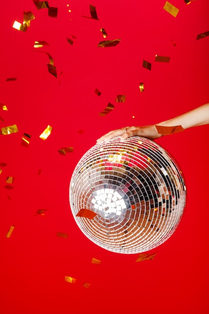 Une main féminine tient une boule disco sur un fond rouge vif et des paillettes d'or volent