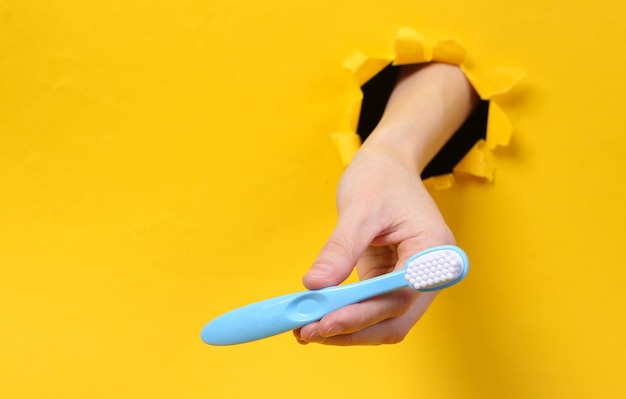 La main féminine tient la brosse à dents à travers le trou déchiré du papier jaune Soins de la cavité buccale Concept art Minimalisme