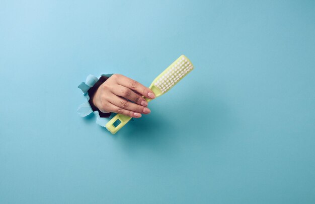 La main féminine tient une brosse en plastique verte sur fond bleu. Une partie du corps sort d'un trou avec des bords déchirés dans un fond de papier