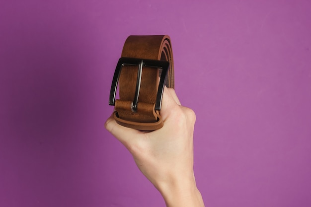 Main féminine tient une ceinture en cuir marron sur fond violet