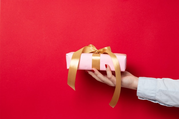 Main féminine tient un emballage cadeau enveloppé dans du papier rose et un noeud de ruban d'or sur rouge