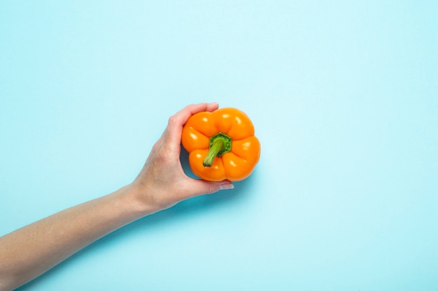 Une main féminine tient un poivron orange sur une surface bleue. Vue de dessus, mise à plat.