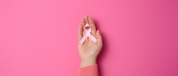 Une main féminine tient un ruban de soie rose en forme de boucle sur fond rose. Symbole de la lutte contre le cancer du sein