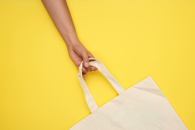 Main féminine tient un sac textile beige vide par les poignées sur jaune, rejet des sacs en plastique, vue du dessus