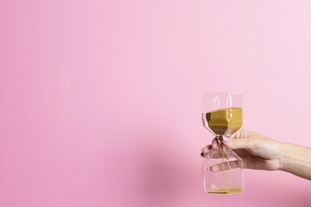 La main féminine tient un verre de sable sur une surface rose