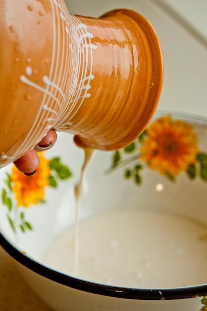Photo main féminine versant du lait d'une casserole en céramique dans un bol en métal peint en émail blanc une cruche en terre cuite brune fermée verticalement