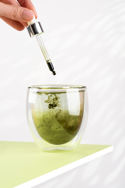 Main féminine verse de la chlorophylle liquide dans un verre d'eau avec un compte-gouttes Verre de chlorophylle liquide sur la table verte Fond blanc Lumière naturelle du soleil Concept de superaliment sain