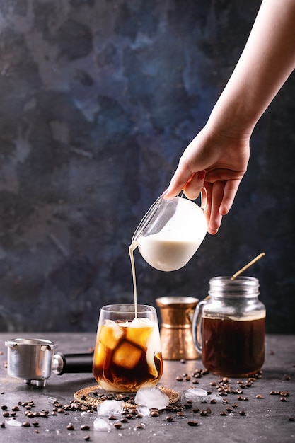 Photo main féminine verser de la crème dans du café glacé sur une surface bleue avec des grains de café et des glaçons autour de