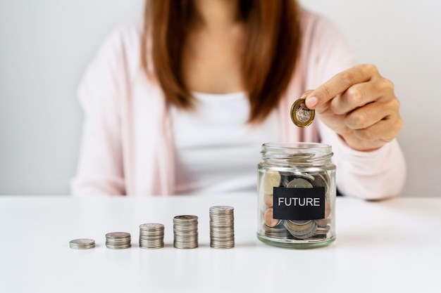 Main De Femme Asiatique Mettant La Pièce Dans Un Bocal En Verre Sur Fond De Tableau Blanc. économiser, Collecter De L'argent Pour L'avenir, Concept D'investissement. Fermer