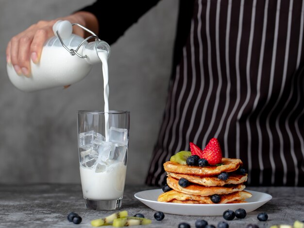 Main de femme au foyer en tablier versant du lait frais de la bouteille à la glace en verre à côté d'une pile de crêpes maison avec garniture de fruits.