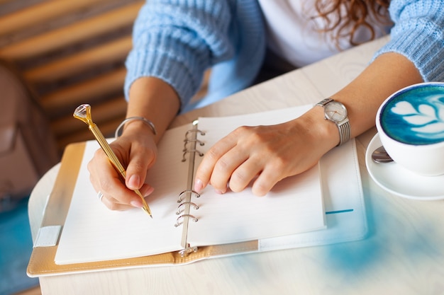 Main de femme bien entretenue tenant un stylo en or et écrire des notes avec un stylo en or dans un cahier tout en buvant du café au lait bleu à côté de la fenêtre. Journaliste indépendant travaillant à domicile. Planification du futur concept. Copier l'espace