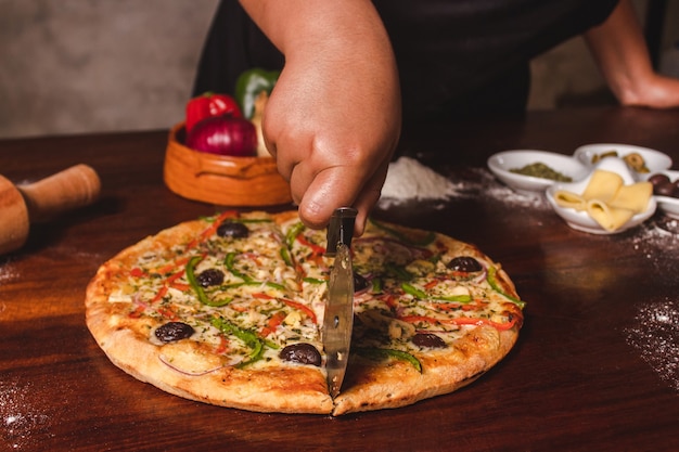 Main de femme coupant la pizza végétarienne en deux sur une table en bois.