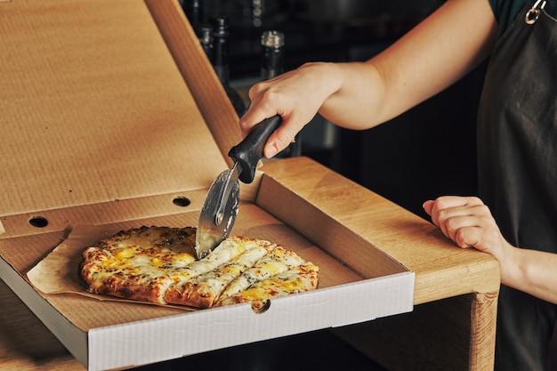 La main de la femme coupe la pizza dans une boîte avec un couteau à rouleau pizza carrée romaine ou pina sur une pâte épaisse