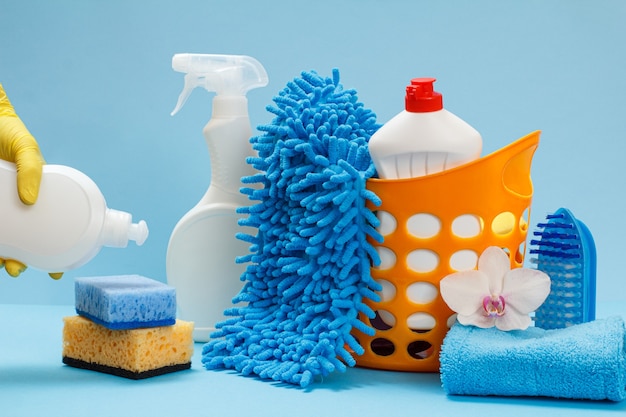 Main de femme dans un gant de protection jaune tenant une bouteille en plastique de liquide vaisselle avec un ensemble de lavage et de nettoyage sur fond bleu.