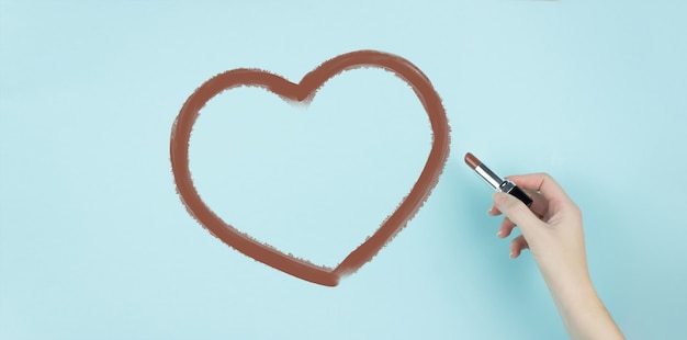 Main de femme dessiner coeur avec rouge à lèvres marron sur fond bleu.