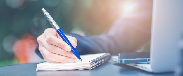 Main de femme écrivant sur un bloc-notes avec un stylo et elle tient un téléphone portable dans le bureau. Bannière Web.