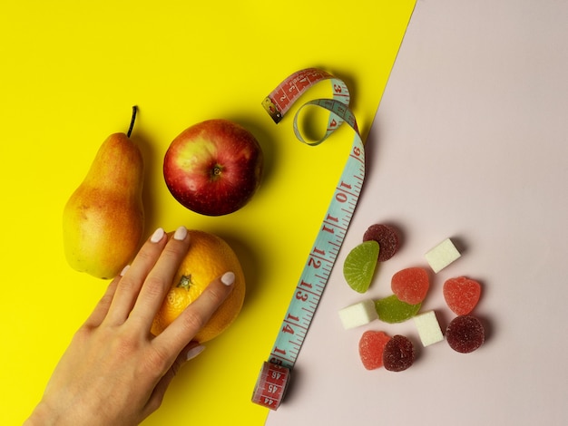 La main d'une femme fait un choix en faveur d'une bonne nutrition. sur fond rose et jaune, des fruits utiles, de l'orange poire pomme et des bonbons nocifs sont disposés. Au milieu se trouve un ruban à mesurer