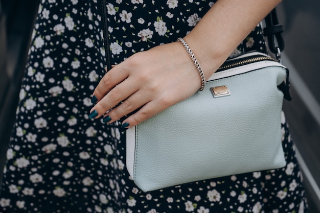 La main de la femme avec manucure et bracelet tient un sac à main. Fille en robe d'été avec un sac à main. Main avec manucure et bijoux tenant un sac à main bleu.