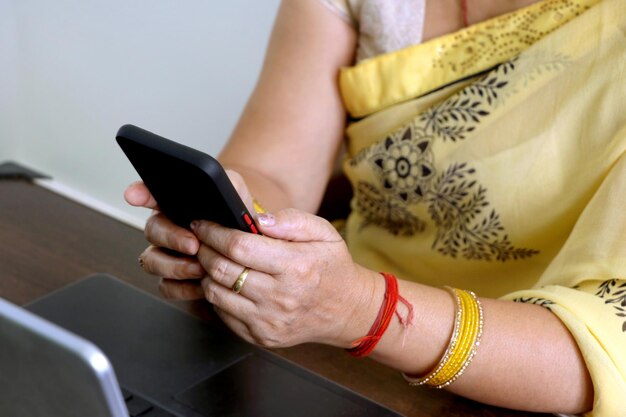 Main d'une femme méconnaissable en tenue indienne Saree utilisant un smartphone tout en travaillant avec un ordinateur portable à la maison