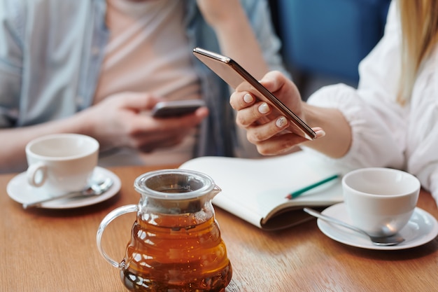 Main de femme millénaire défilement dans le smartphone sur la table tout en ayant une tasse de thé au café avec son petit ami ou camarade de groupe