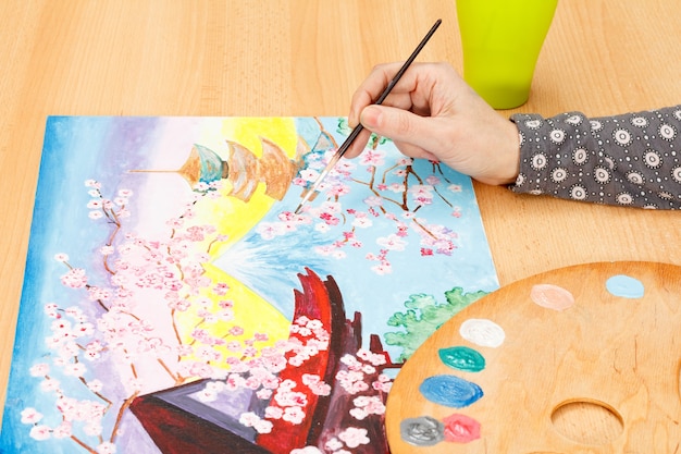 Main de femme peignant un paysage japonais pittoresque à l'intérieur sur le bureau en bois avec des peintures et un pinceau