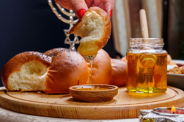 La main d'une femme plonge un morceau de challah dans du miel en l'honneur de la célébration de Rosh Hashanah près du miel et de la challah et de la nourriture traditionnelle