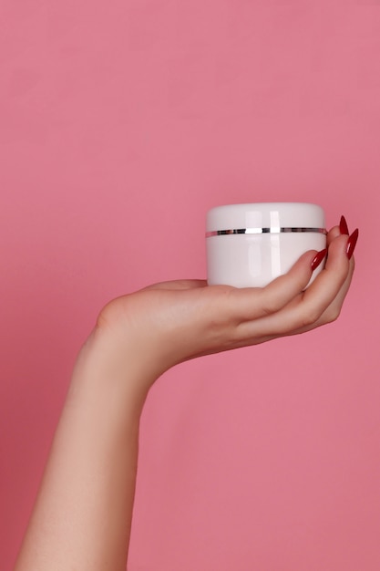 Main de femme avec un pot de crème sur fond rose