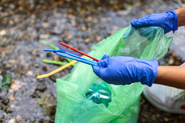 Main de femme ramassant des pailles en plastique dans des sacs à ordures tout en nettoyant une petite rivière