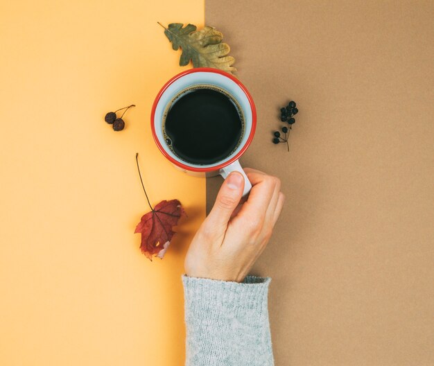 Une main de femme avec une tasse de café et des feuilles séchées.