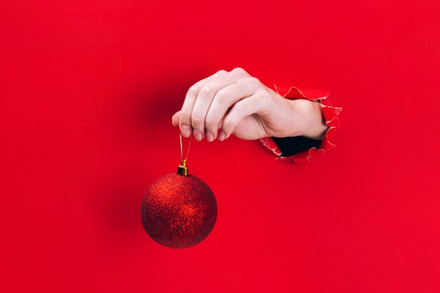 Main de femme tenant une boule d'arbre de Noël rouge scintillant à travers un trou en rouge.